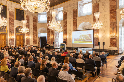 Blick in den Rittersaal. An der Decke hängen Kronleuchter, das Publikum sitzt in Stuhlreihen vor der Bühne und ist von hinten zu sehen. Auf der Bühne steht Professor Fetzer, neben ihm ist eine Leinwand zu sehen.