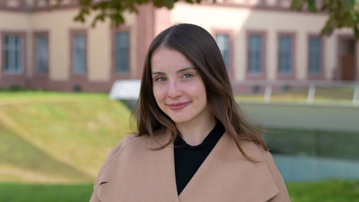 Eine lächelnde Person trägt ein schwarzes Top sowie einen hellbraunen Mantel und steht auf der Mensawiese. Die Person heißt Anna Valerie Smeljanski.