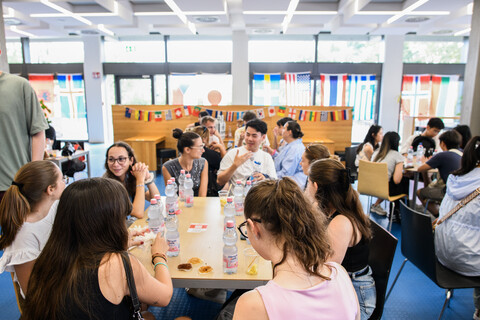 Studierende sitzen an einem Tisch in der Mensa und unterhalten sich. Im Vordergrund zwei Studentinnen, die von hinten zu sehen sind.