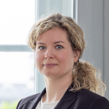 Nora Zybura hat ihre blonden lockigen Haare zu einem Zopf gebunden. Sie trägt Creolen-Ohrringe und einen schwarzen Blazer.