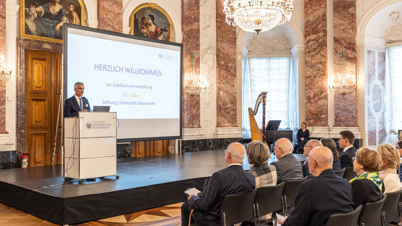 Mehrere Personen sitzen auf Stühlen und hören einer Person zu, die auf einer Bühne vor einem Pult steht. Diese Person ist Prof. Dr. Thomas Fetzer, Rektor der Universität Mannheim.