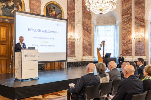 Mehrere Personen sitzen auf Stühlen und hören einer Person zu, die auf einer Bühne vor einem Pult steht. Diese Person ist Prof. Dr. Thomas Fetzer, Rektor der Universität Mannheim.
