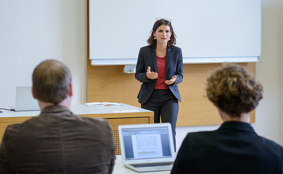 Eine Dozentin steht vor einer Tafel und hält einen Vortrag. Man sieht zwei Teilnehmer von hinten.