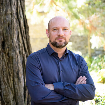  Prof. Ulrich Wagner. Er steht freundlich dreinblickend in blauem Hemd vor einem Baum.