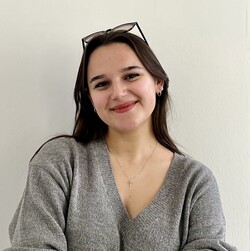 Radina Slavova steht im Innenraum des Schlosses, hat die Arme vor der Brust verschränkt und lacht in die Kamera. Sie trägt einen grauen Pullover mit V-Ausschnitt und hat die Sonnenbrille ins braune, lange Haar zurückgeschoben.