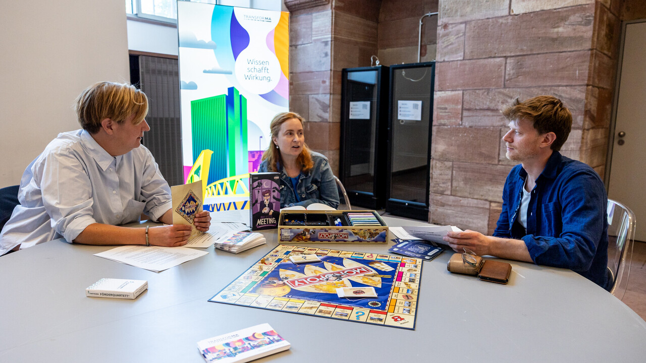 Drei Personen sitzen in den Katakomben an einem Tisch, auf dem der Spielplan des Spiels "Monopoly" ausgebreitet ist. Es befinden sich noch weitere Gegenstände auf dem Tisch, darunter die Spiele "Förderquartett" und "Das letzte Meeting". Im Hintergrund ein Plakat des Projekt "TransforMA".