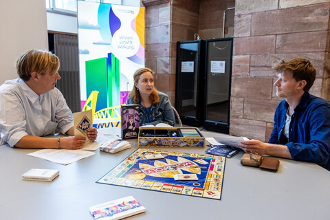 Drei Personen sitzen in den Katakomben an einem Tisch, auf dem der Spielplan des Spiels "Monopoly" ausgebreitet ist. Es befinden sich noch weitere Gegenstände auf dem Tisch, darunter die Spiele "Förderquartett" und "Das letzte Meeting". Im Hintergrund ein Plakat des Projekt "TransforMA".