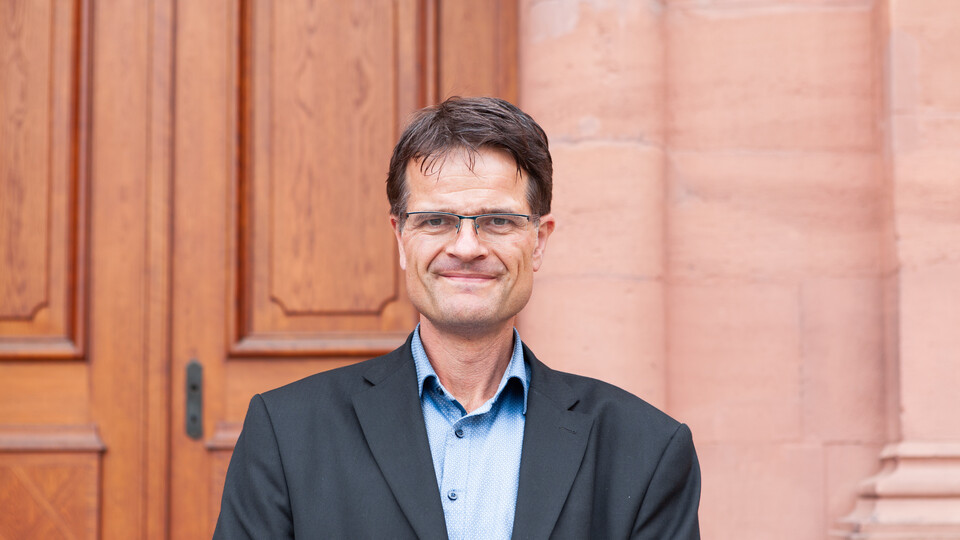 Portrait von Bernward Gesang. Er trägt ein hellblaues Hemd sowie einen dunkelgrauen Blazer und steht lächelnd vor dem Schloss. 
