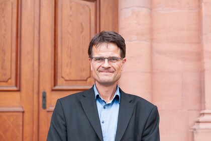 Prof. Dr. Gesang trägt einen schwarzen Anzug mit hellblauem Hemd und steht vor einer Eingangstür des Universitätsgebäudes.