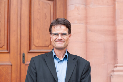 Portrait von Bernward Gesang. Er trägt ein hellblaues Hemd sowie einen dunkelgrauen Blazer und steht lächelnd vor dem Schloss. 