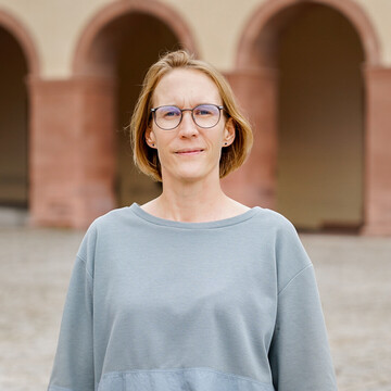 Portraitaufnahme von Regine Zeller im Ehrenhof des Mannheimer Schlosses, Regine Zeller hat halblange braune Haare, trägt eine Brille und einen grauen Pullover