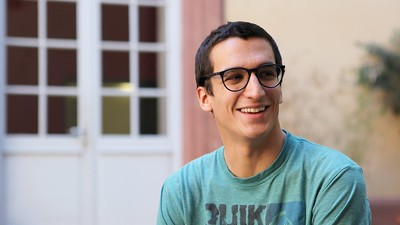 Eine lächelnde Person trägt ein türkisfarbenes T-Shirt und sitzt in einem Innenhof. Die Person heißt Santiago Rey-Sanchez Parodi.