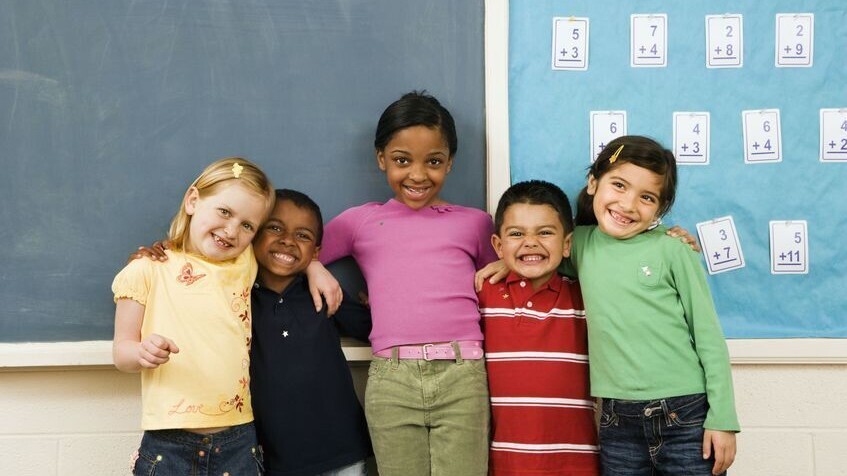 vier diverse Grundschulkinder stehen in einem Klassenraum vor einer Tafel. Sie umarmen sich und lachen.