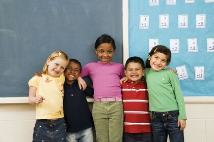 vier diverse Grundschulkinder stehen in einem Klassenraum vor einer Tafel. Sie umarmen sich und lachen.