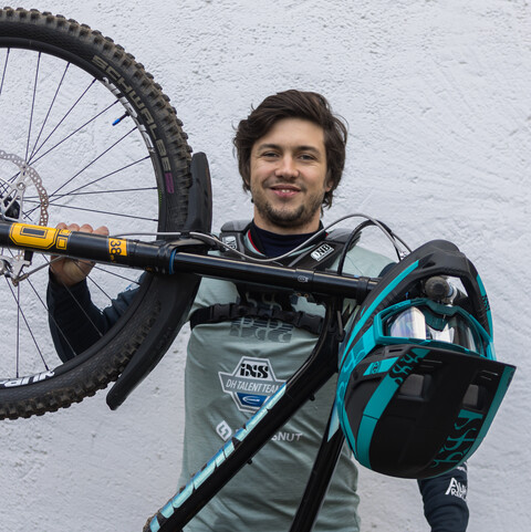 Eine Person trägt schwarze und graue Sportbekleidung und steht vor einer weißen Wand. Die Person hält ein Mountainbike, welches nur mir dem Hinterrad auf dem Boden steht. Die Person heißt Jannik Abbou.