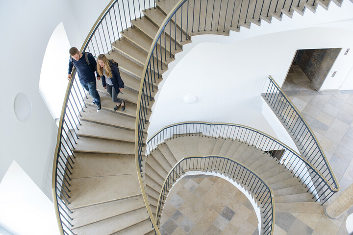 Zwei Studierende laufen eine Wendeltreppe aus Stein in der Uni hinunter
