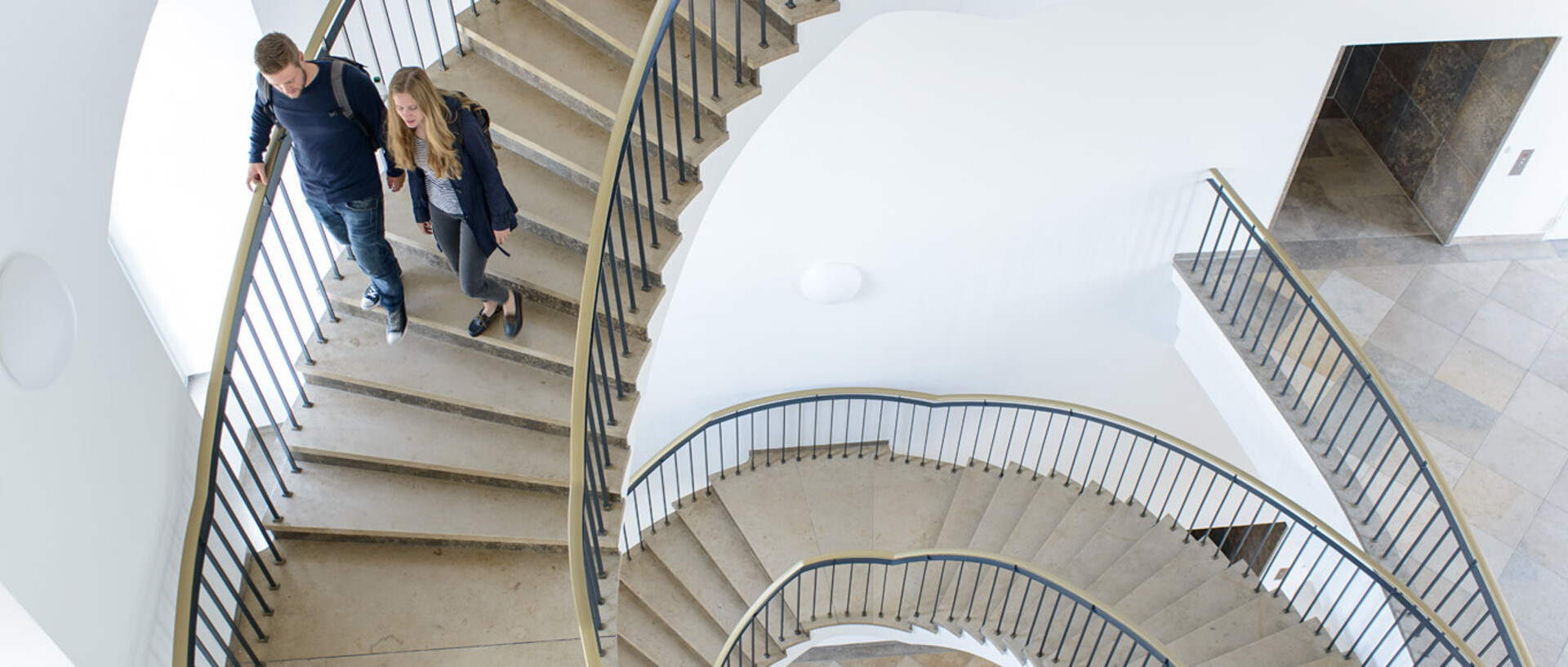 Zwei Studierende laufen eine Wendeltreppe aus Stein in der Uni hinunter