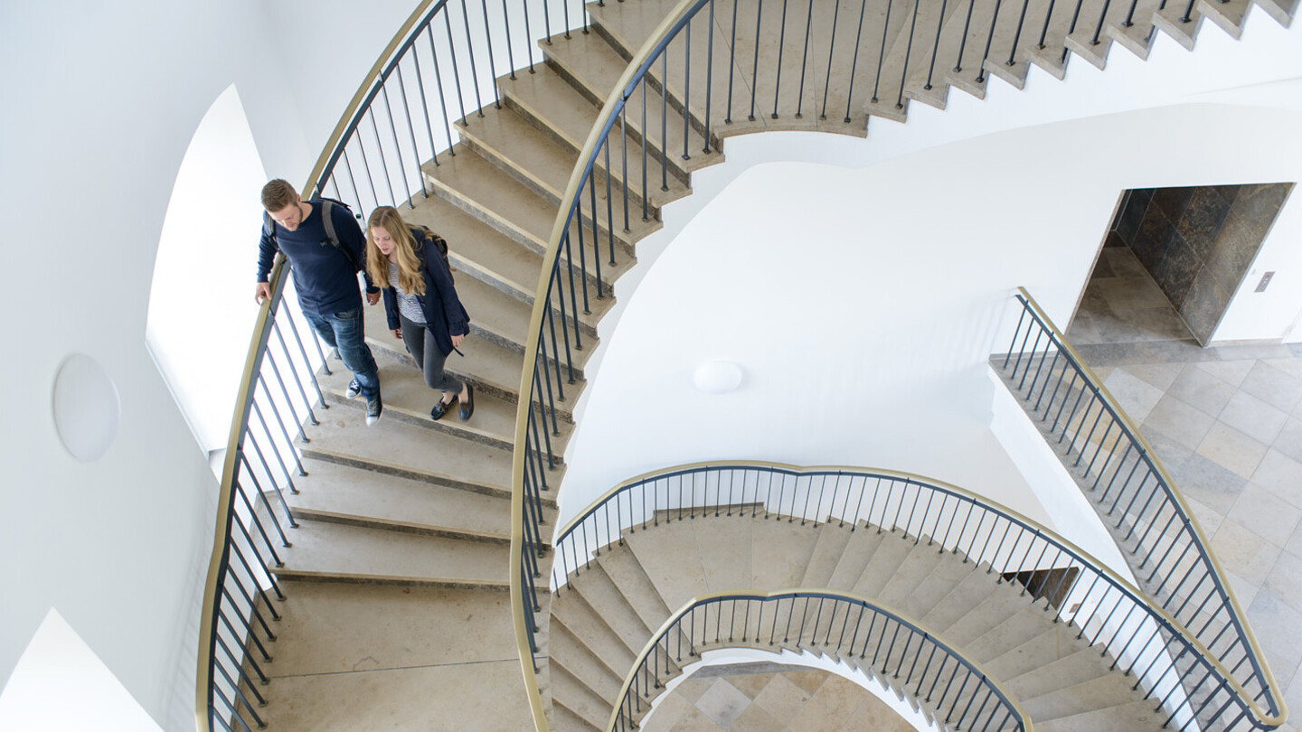 Zwei Studierende laufen eine Wendeltreppe aus Stein in der Uni hinunter