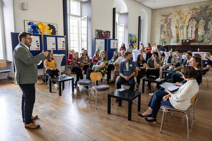eine Person steht in einem festlichen Saal der Universität und spricht vor einer Gruppe sitzender Personen