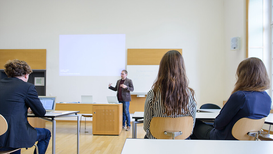 Ein Dozent steht vorne und hält einen Vortrag. Ein Beamer ist aufgebaut und man sieht Studierende von hinten.