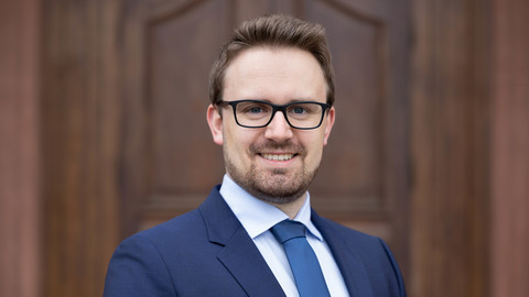 Alexander Rupertus. He is wearing a blue suit and is standing in front of brown door.