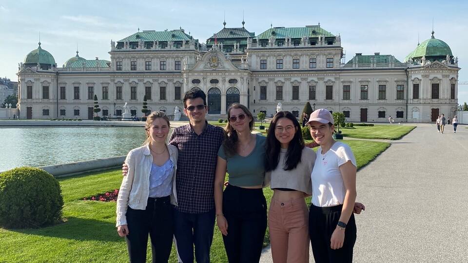 Fünf lächelnde Personen stehen nebeneinander in einem Schlossgarten vor einem Schloss.