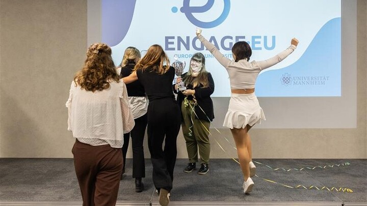 Mehrere Personen stehen auf einer Bühne und feiern; eine Person hält einen Pokal. Im Hintergrund ist das ENGAGE.EU-Logo an die Wand projiziert.