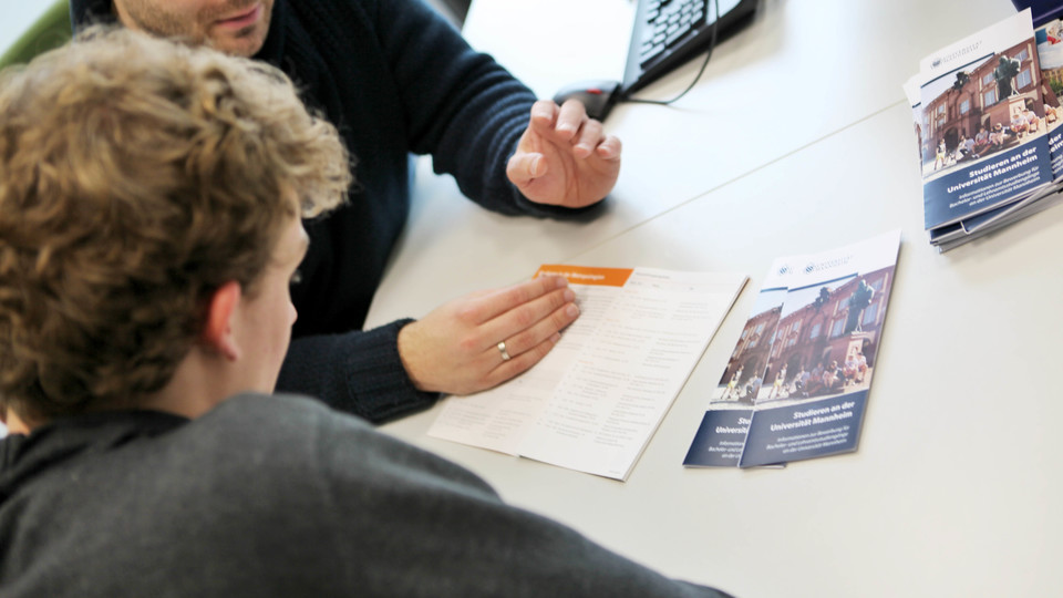 Ein Mitarbeiter berät einen Studenten. Beide blicken in eine Broschüre vor ihnen auf dem Tisch.