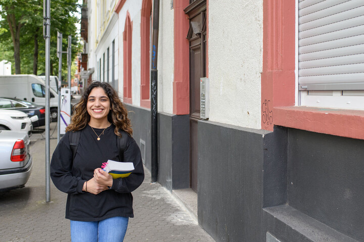 Eine Studierende lächelt und läuft mit einem Buch in der Hand durch eine Straße in Mannheim.