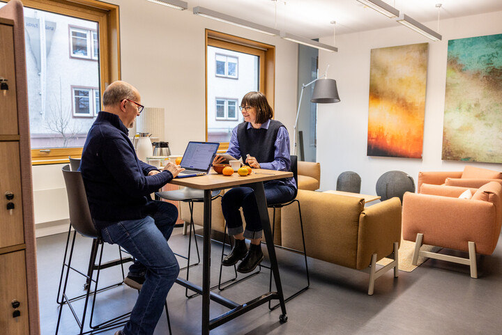 Das Bild zeigt zwei Fellows. Sie sitzen auf hohen Barhockern an einem Stehtisch gegenüber und unterhalten sich. Sie lachen.