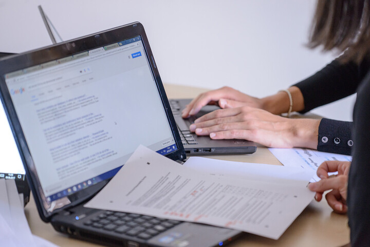 Zwei Personen sitzen an einem Tisch und arbeiten an ihren Laptops. Wir sehen nur ihre Hände. Auf einem Laptop liegen Aufgabenblätter mit Notizen.