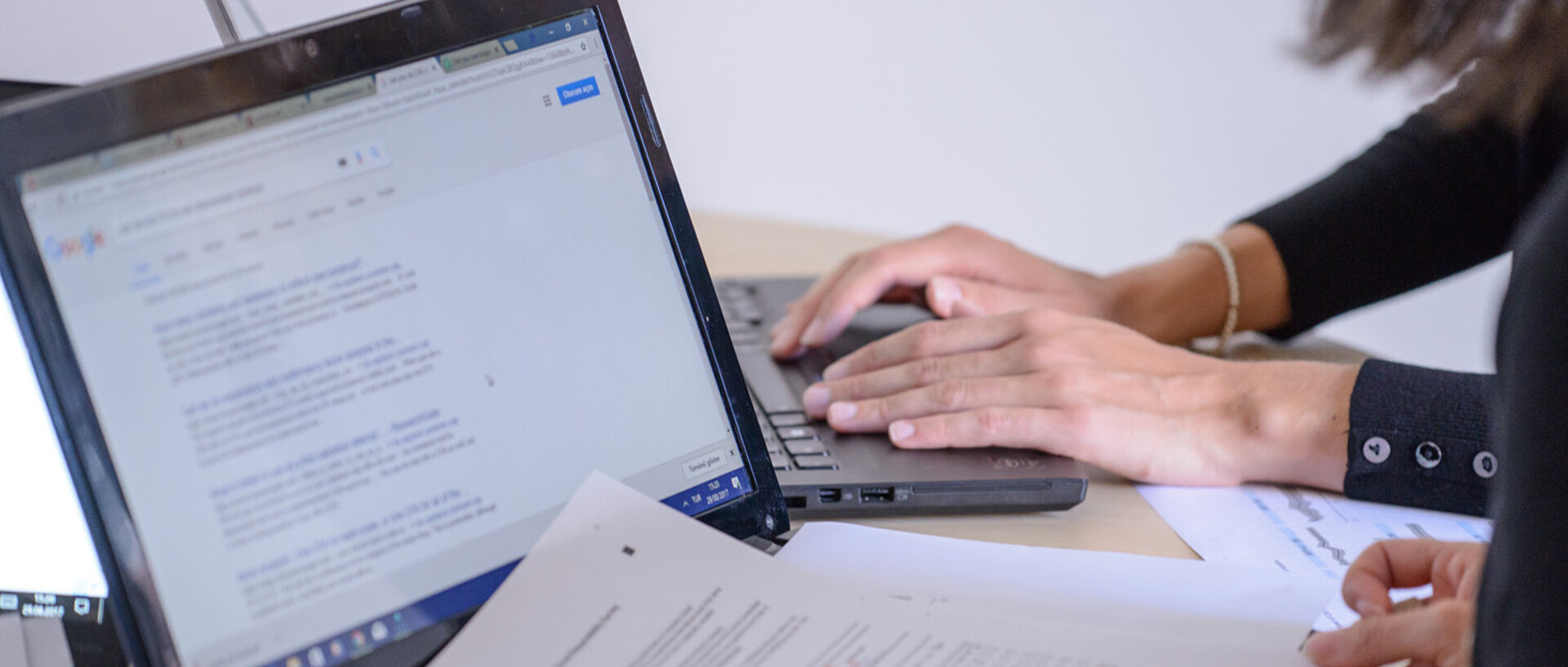 Zwei Personen sitzen an einem Tisch und arbeiten an ihren Laptops. Wir sehen nur ihre Hände. Auf einem Laptop liegen Aufgabenblätter mit Notizen.