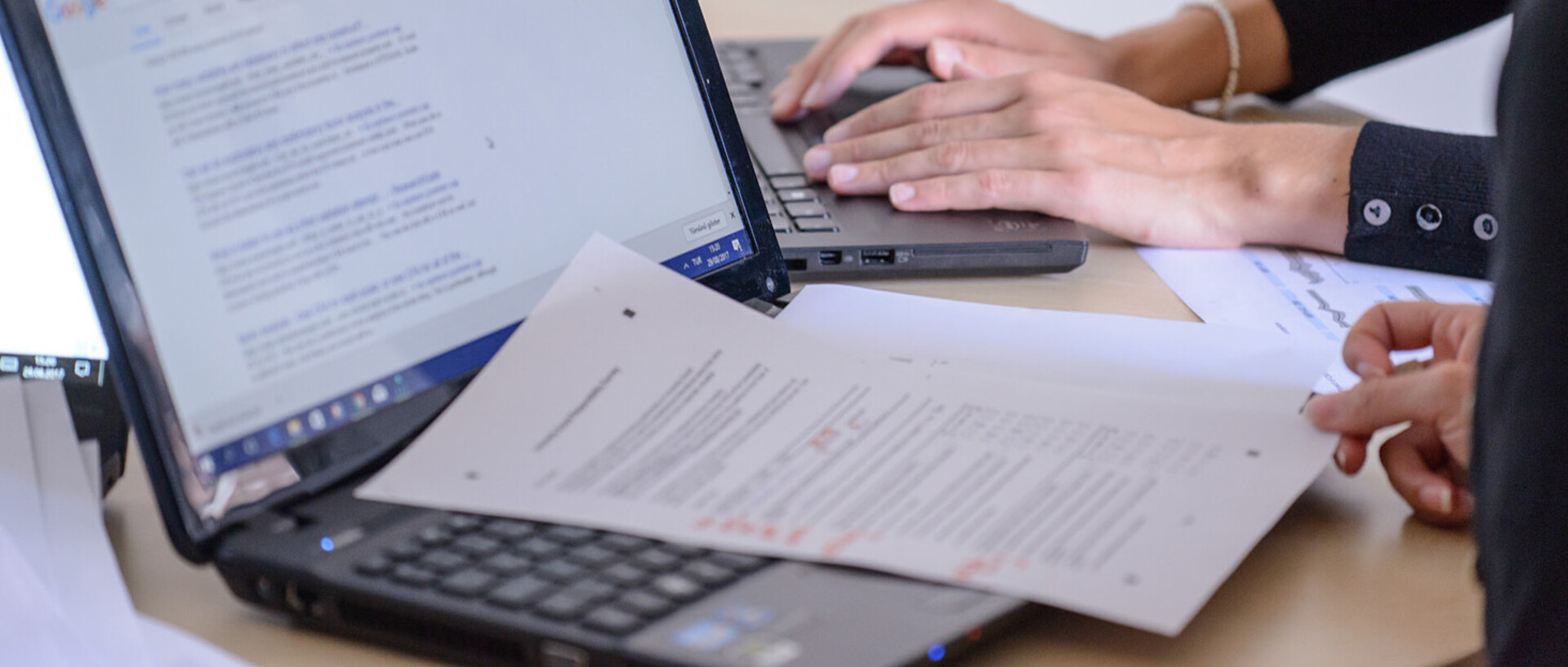 Zwei Personen sitzen an einem Tisch und arbeiten an ihren Laptops. Wir sehen nur ihre Hände. Auf einem Laptop liegen Aufgabenblätter mit Notizen.