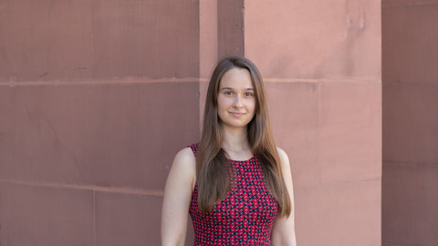 Barbara Kreis. She is wearing a red sleeveless dress and is standing in front of a stonewall.