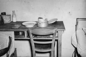 schwarz-weiß Aufnahme eines damaligen Wohnbunkers der Universität Mannheim. Kleiner Holztisch mit Geschirr und Töpfen darauf. Davor ein Holztisch und daneben das Bett. 