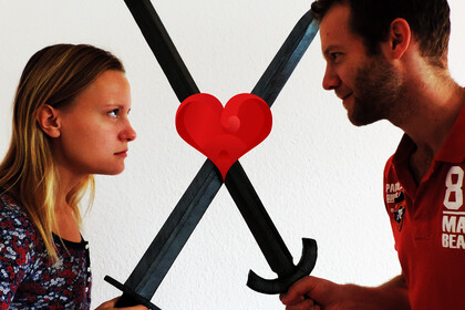 Ein Mann und eine Frau mit schwarzen Schwertern stehen sich gegenüber. Sie schauen sich tief in die Augen. Die Schwerter zeigen jeweils schräg nach oben, an der Stelle, an der sie sich kreuzen befindet sich ein rotes Herz.. 