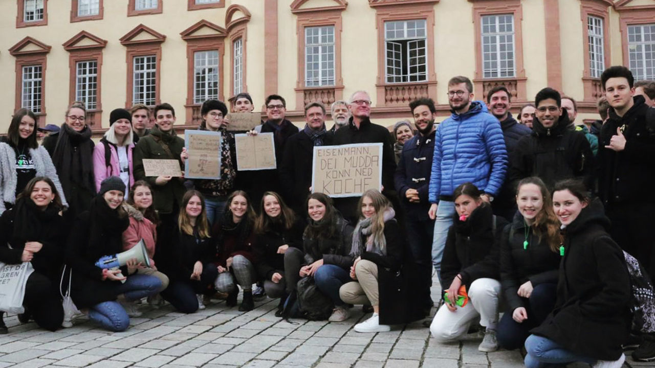 Eine Gruppe aus Studierenden steht vor dem Mannheimer Schloss und demonstriert. Sie haben Pappschilder und ein Megaphon dabei.