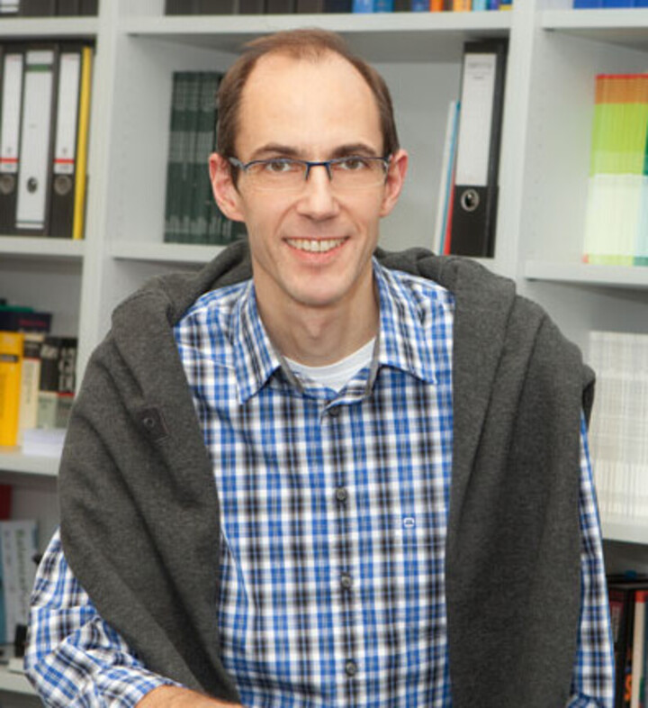 Portrait von Thorsten Meiser. Er trägt ein blau-weiß karriertes Hemd, sowie einen grauen Pullover über seinen Schultern und sitzt vor einem Regal. 