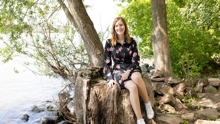 Eine Studentin mit schwarzem Kleid und pinken Rosen sitzt auf einem Baumstumpf am Wasser.