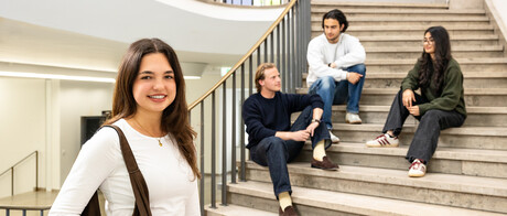 Eine Studentin steht im Treppenhaus des Schlosses. Auf der Treppe unterhalten sich drei Studierende.