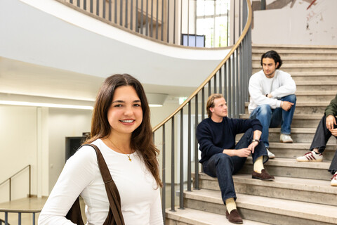 Eine Studentin steht im Treppenhaus des Schlosses. Auf der Treppe unterhalten sich drei Studierende.