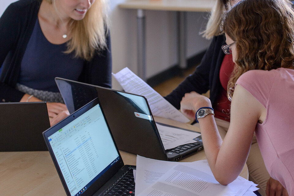 Drei Studentinnen sitzen vor ihren Laptops. Sie haben sich einander zugewandt und arbeiten an Aufgabenblättern.