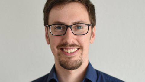 Joscha Krug. He is wearing a dark blue shirt and is standing in front of a beige background.