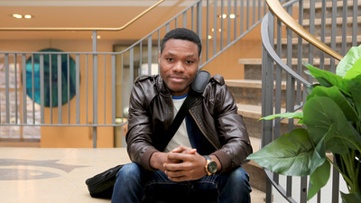 Eine Person trägt ein weißes T-Shirt, eine braune Lederjacke sowie eine dunkelblaue Jeans und sitzt auf einer Treppe. Die Person heißt Louis Anyanwu.