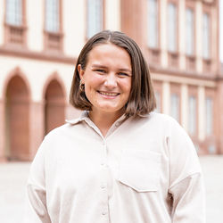 Ein Porträt von Sophie Rudolph, die in die Kamera lächelt. Sie trägt schulterlange braune Haare und ein beigefarbenes Hemd. Ihre Hände sind lässig in die blauen Jeanstaschen gesteckt.