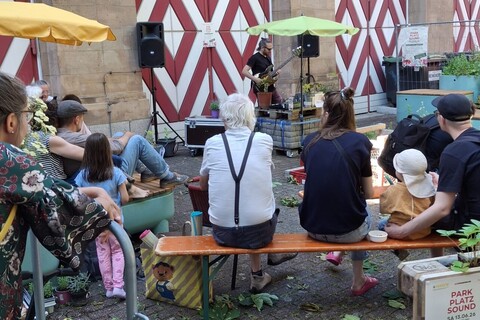 Mann mit Gitarre, mehrere sitzende Leute die zum Mann mit Gitarre schauen vor einem Haus mit rot-weiß-gestreiften HolztürenMann mit Gitarre, mehrere sitzende Leute die zum Mann mit Gitarre schauen vor einem Haus mit rot-weiß-gestreiften Holztüren