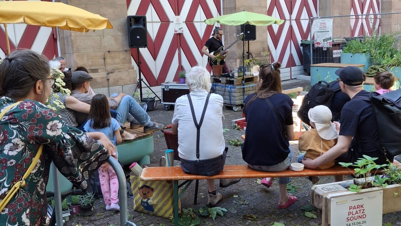 Mann mit Gitarre, mehrere sitzende Leute die zum Mann mit Gitarre schauen vor einem Haus mit rot-weiß-gestreiften HolztürenMann mit Gitarre, mehrere sitzende Leute die zum Mann mit Gitarre schauen vor einem Haus mit rot-weiß-gestreiften Holztüren