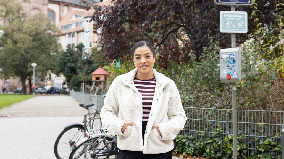 Eine Studentin in der Mannheimer Innenstadt. Im Hintergrund sind angeschlossene Fahrräder und ein Straßenschild mit der Aufschrift "Schillerplatz".