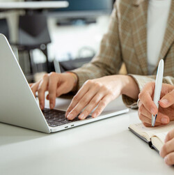 Eine Frau tippt am Laptop, ein Mann sitzt daneben mit Block und Stift.