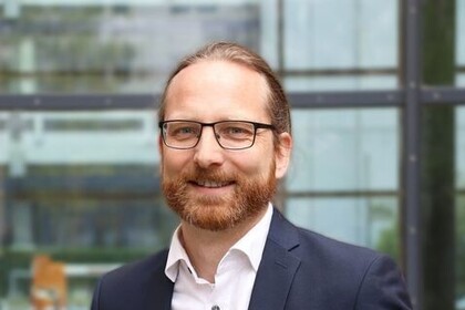 Heiner Stuckenschmidt hat rötlich-braune, zu einem Zopf gebundene Haare. Er trägt eine eckige Brille, ein weißes Hemd und ein dunkles Jacket